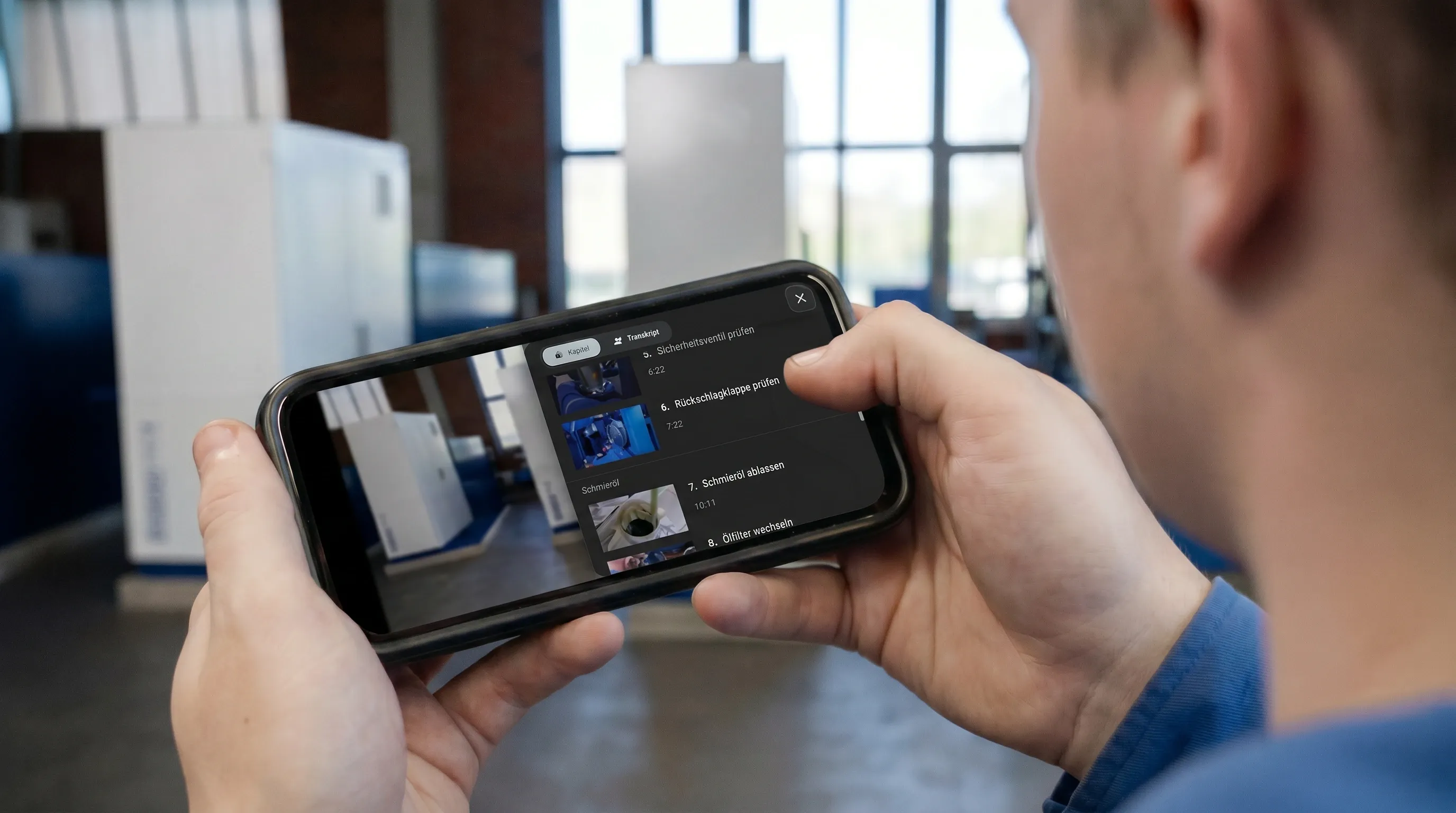 Servicetechniker hält ein Smartphone mit einer interaktiven Videoanleitung, sichtbar ist die Kapitelliste mit Wartungsschritten wie Sicherheitsventil prüfen und Schmieröl ablassen.